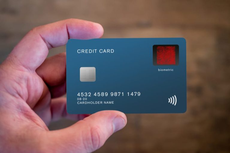 Sebuah tangan memegang kartu kredit modern berwarna biru tua. Kartu ini dilengkapi chip, panel fingerprint merah bertuliskan "biometric", dan simbol nirkontak di bagian kanan bawah