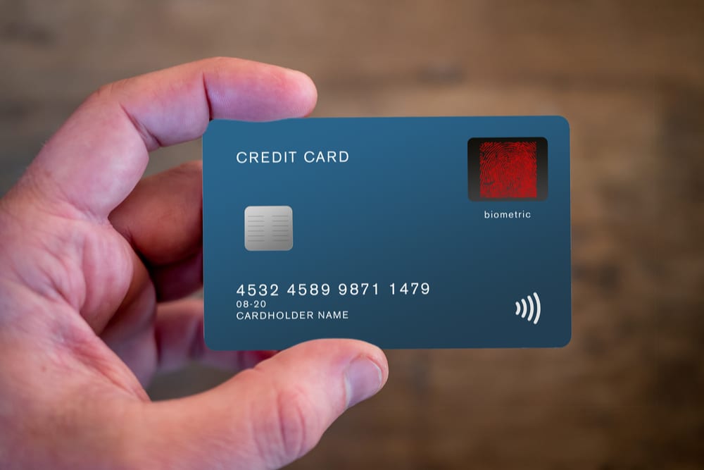 Sebuah tangan memegang kartu kredit modern berwarna biru tua. Kartu ini dilengkapi chip, panel fingerprint merah bertuliskan "biometric", dan simbol nirkontak di bagian kanan bawah